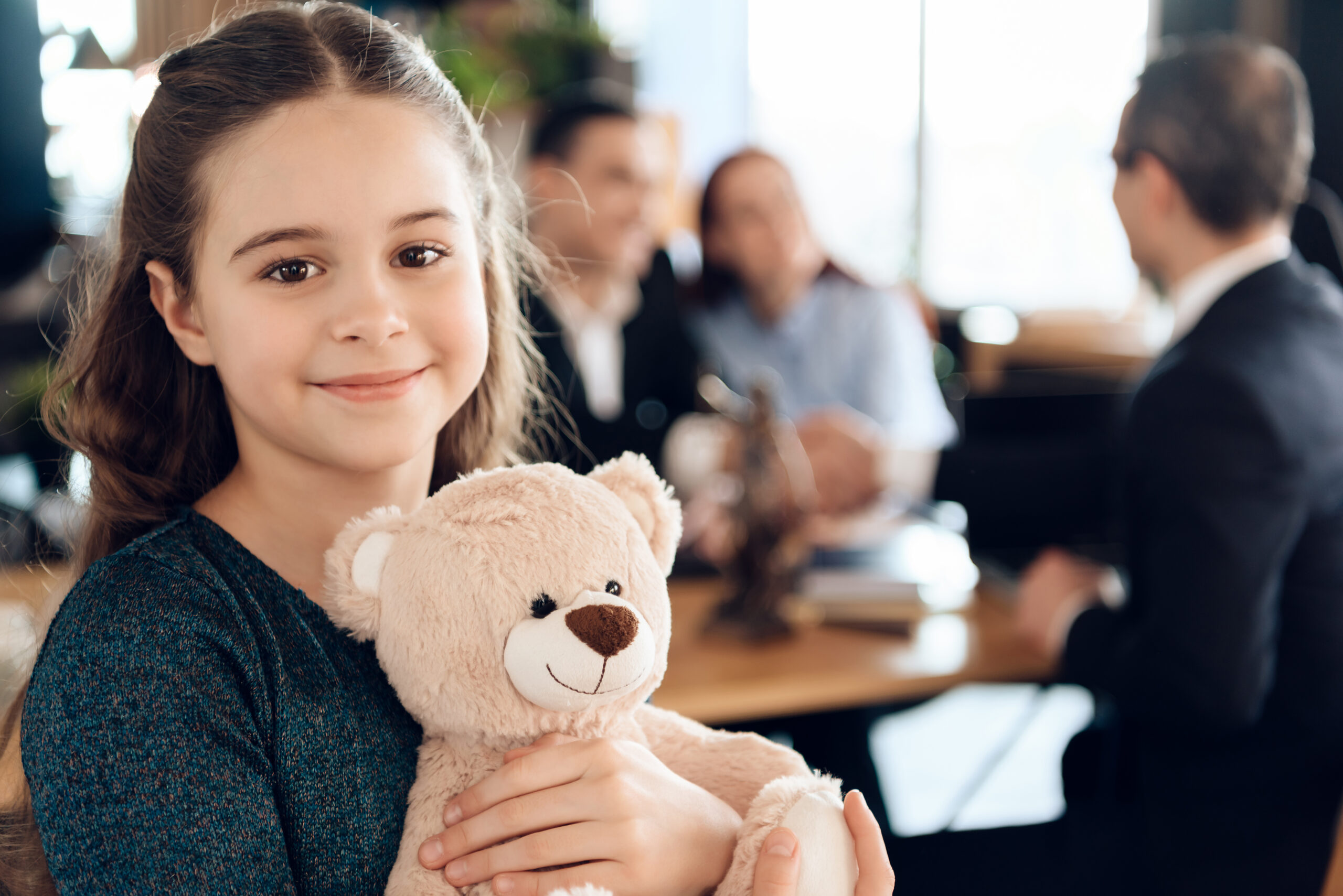 little girl holding a teddy bear in an office while parents speak to a lawyer in the background