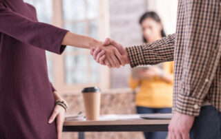 Male hand and female hand shaking hands in office with women and desk in the background